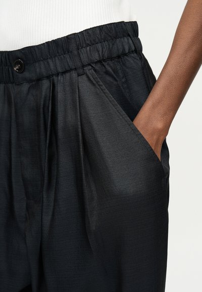 Κοντινή λήψη σκούρων, φαρδιών παντελονιών με ελαστική ζώνη μέσης και κουμπί, που φοριούνται με το χέρι να τοποθετείται άνετα στην εμπρόσθια τσέπη.