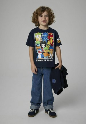 Camiseta marina con personajes de dibujos animados coloridos y el texto "¡TROPEZANDO TODO EL DÍA!". Combinada con jeans azules y zapatillas marinas con acentos blancos.