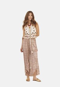 Chaleco de ganchillo en color crema con patrones geométricos, combinado con pantalones holgados rosas con estampado y aberturas laterales. Modelo lleva sandalias doradas.