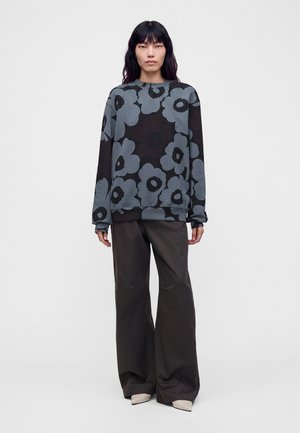 Femme portant un sweat-shirt imprimé floral noir et gris, un pantalon large noir et des chaussures claires, debout devant un fond blanc uni.