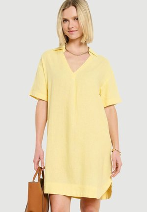 Žena v voľných krátkych žltých šatách drží hnedú kabelku a stojí na neutrálom pozadí.