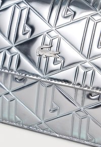 Metalen zilveren leren tas met geometrisch patroon, gestikte randen en klein alligatorlogo in het midden.