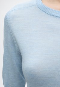 Ανοιχτό μπλε πλεκτό πουλόβερ που φοράει ένα άτομο, εμφανίζοντας τον ώμο και το πάνω μέρος του στήθους με στρογγυλή λαιμόκοψη.