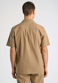 Camisa de manga corta con textura en tejido marrón claro, con un cuello relajado y detalle de pliegue en la espalda, mostrada desde atrás.