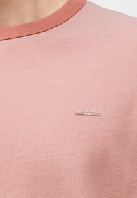 Κοντινό πλάνο ενός ροζ μπλουζιού με υφή και στρογγυλή λαιμόκοψη, που φέρει ένα μικρό μπεζ λογότυπο "JACK & JONES" στην αριστερή πλευρά του στήθους.