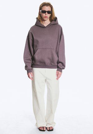 Personne aux longs cheveux blonds portant des lunettes de soleil noires, un sweat à capuche violet ample, un pantalon blanc large et des sandales noires ouvertes, debout sur fond blanc.