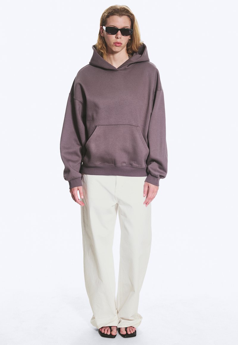 Personne aux longs cheveux blonds portant des lunettes de soleil noires, un sweat à capuche violet ample, un pantalon blanc large et des sandales noires ouvertes, debout sur fond blanc.