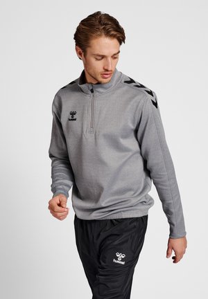 Grijze half-zip pullover met een textuurfinish, voorzien van een zwart logo en chevronaccenten op de schouders, gecombineerd met zwarte losvallende broek.