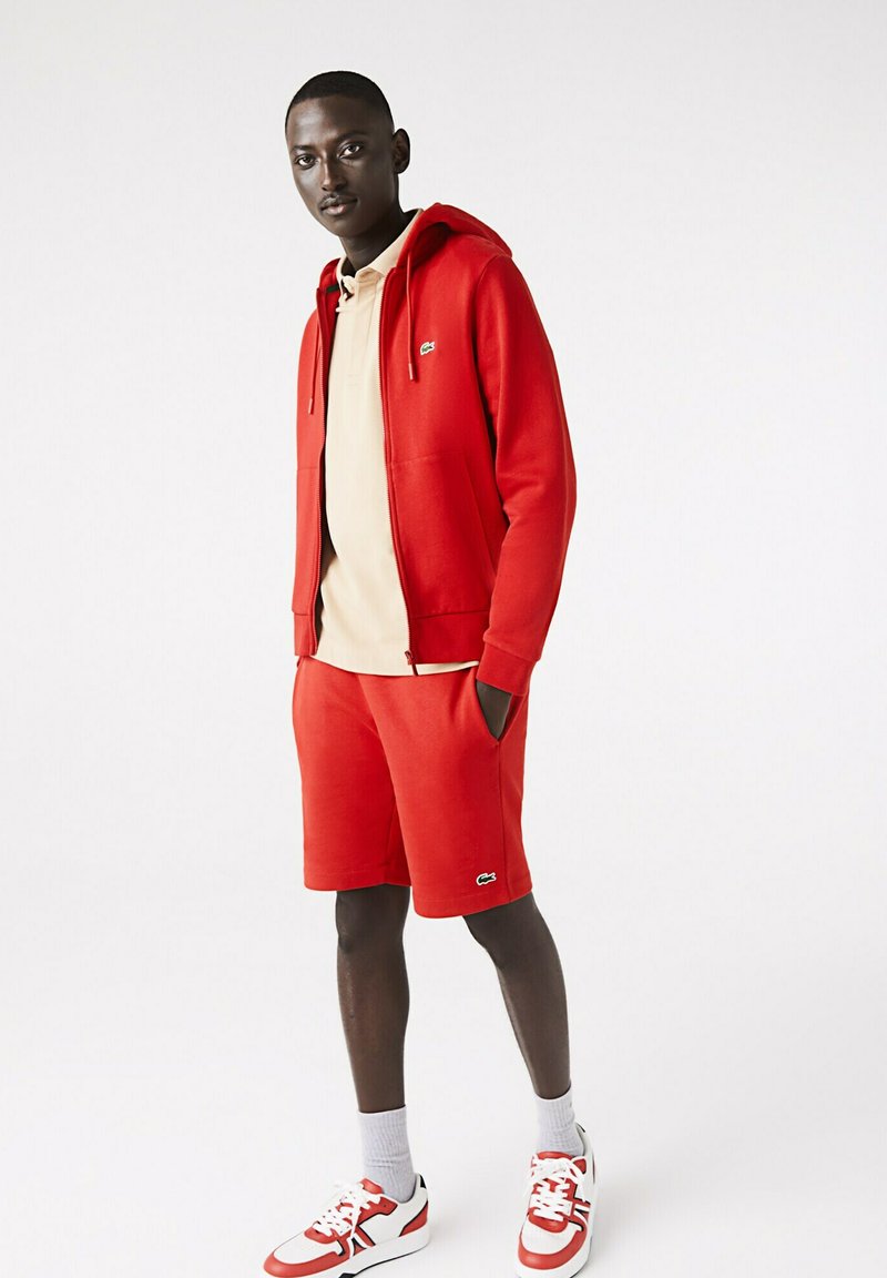 Rote Zip-Hoodie mit Taschen, kombiniert mit passenden roten Shorts. Beige Poloshirt darunter. Rote und weiße Sneakers, weiße Socken. Lacoste-Logo sichtbar.