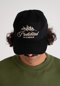 Svart bomullskap med "Forbudt" og "10119 BERLIN" i hvit brodering, pyntet med et bladdesign. Myk tekstur og buet skygge.