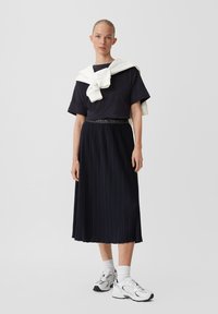 Gonna plissettata blu marina abbinata a una maglietta nera a maniche corte e a un maglione bianco appeso al collo; completata con scarpe da ginnastica bianche.