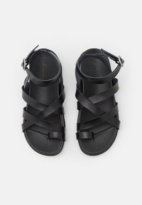 Schwarze Ledersandalen mit mehreren kreuzenden Riemen, einem Knöchelriemen mit einer runden Metall-Schnalle und einer flachen Sohle mit strukturierter Oberfläche.