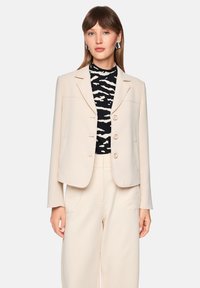 Light beige blazer with a tailored fit, two front pockets, and round buttons. Worn over a black and white striped top. Smooth texture.