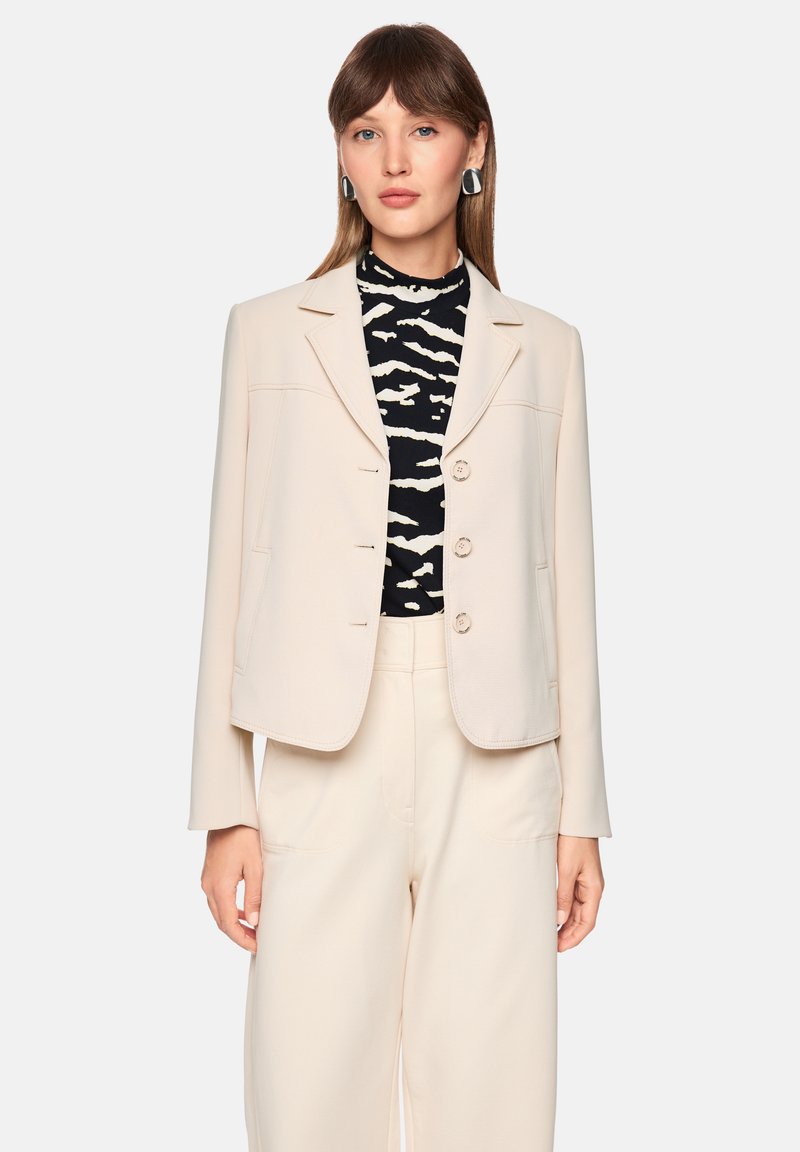 Light beige blazer with a tailored fit, two front pockets, and round buttons. Worn over a black and white striped top. Smooth texture.