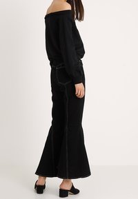 Top negro sin hombros combinado con vaqueros de campana negros con detalles de costura blanca. El modelo lleva mules negros de tacón.