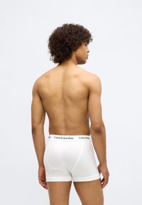 White cotton boxer briefs with a contrasting black waistband featuring "Calvin Klein" logo. Model shows the garment's fit from the back.