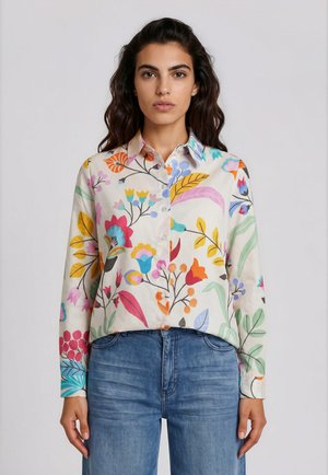 Frau trägt ein langärmliges Hemd mit bunten Blumenmustern und blaue Jeans, steht vor einem schlichten hellen Hintergrund.