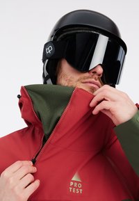 Rote wasserdichte Jacke mit grünem Futter, Reißverschlusskragen und gut sichtbarem Logo. Getragen mit einem schwarzen Helm und einer Schutzbrille.