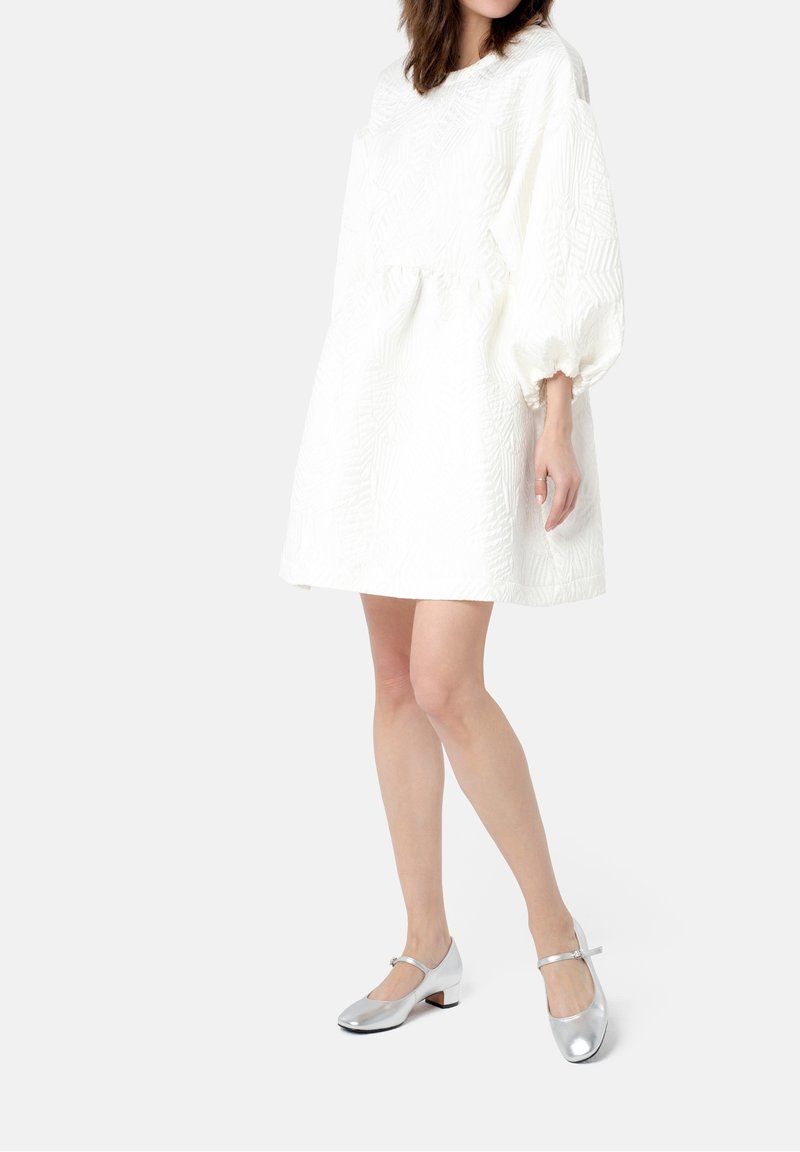 Robe blanche texturée à coupe ample avec manches bouffantes, associée à des chaussures Mary Jane argentées avec un petit talon et une bride.