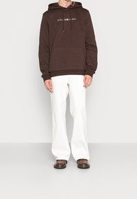 Bruine hoodie met witte tekst, voorzak en trekkoorden. Draag het met witte wijde pantalons en bruine schoenen. Eenvoudig en casual ontwerp.