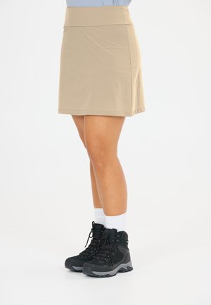 Persoon die een beige knielange rok, lichtblauw shirt, witte sokken en zwarte wandelschoenen draagt, staand op een witte achtergrond.