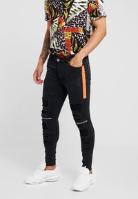 Jeans ajustados negros con una franja lateral naranja, detalles desgastados y acentos de cremallera. Combinados con una camisa de estampado gráfico multicolor y zapatillas blancas.