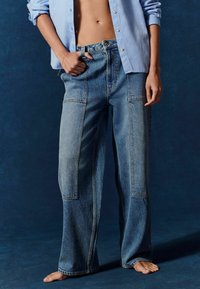 Széles lábú kék farmernadrág, elhalványult foltokkal, magas derékkal, látható varrásokkal és nagy elülső zsebekkel, világoskék inggel párosítva.