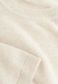 Pull en tricot beige avec un motif texturé, doté d'un col et de poignets côtelés, mettant en valeur un tissu doux et une coupe ample.