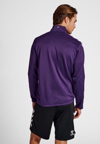 Fioletowa sportowa kurtka z wysokim kołnierzem i długimi rękawami, wykonana z gładkiego materiału. Noszona z czarnymi szortami z logo i bocznymi akcentami.