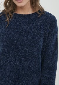 Femme portant un pull en chenille texturé bleu marine avec un col rond et des manches longues, sur un fond uni.