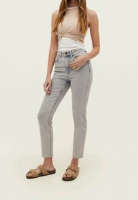 Nuori nainen, jolla on päällään solmittu hihaton beige toppi, vaaleansiniset korkeavyötäröiset farkut ja vaaleat solkisaappaat, seisoo yksinkertaista taustaa vasten.