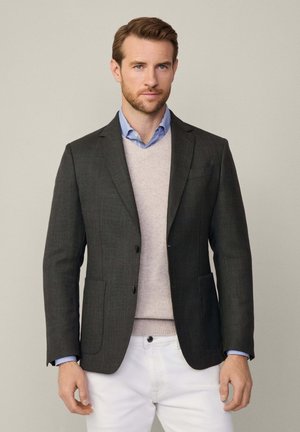 Grau melierter Blazer mit Reverskragen, zwei Vorderpockets, taillierte Passform, über einem hellen Pullover und blauem Hemd getragen, kombiniert mit weißen Hosen.