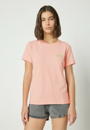 Rožinė trumpomis rankovėmis marškinėliai iš tekstūruotos medžiagos, su mažu žaliu logo ant krūtinės, derinami su pilkais džinsiniais šortais.