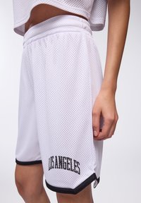 Persona che indossa pantaloni corti da basket bianchi in rete con bordi neri e testo "LOS ANGELES", abbinati a un top corto coordinato.