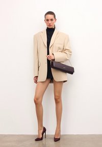 Bežový oversize blejzer s jedným gombíkom, nosený cez čierne rolákové šaty. Tmavo fialová kožená kabelka a zodpovedajúce vysoké podpätky. Hladká textúra.