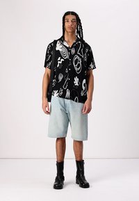 Camisa negra de manga corta con patrones abstractos blancos, combinada con pantalones cortos de denim azul claro y botas negras hasta el tobillo. Bolso sencillo colgado del hombro.