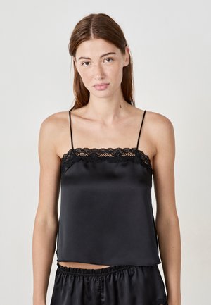 Femme aux longs cheveux bruns portant un débardeur satin noir avec des dentelles et un short satin noir assorti, sur un fond clair et uni.
