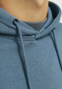 Blå sweatshirt med tekstureret stof, der har en snoretrækshætte og forstærket syning ved halsen og ærmerne.
