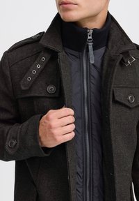 Abrigo de espiga gris oscuro con bolsillos con botón, hombreras y una capa interior negra con cremallera. Presenta un cuello alzado y texturas contrastantes.