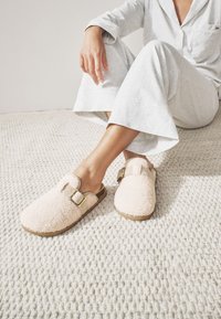 Różowe, puszyste kapcie o miękkiej fakturze, z klamrą i beżową podeszwą, noszone z jasnoszarym strojem do domu, na teksturowanym kremowym dywanie.