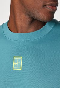 Sudadera de algodón color teal con cuello redondo acanalado, logo bordado en amarillo en el pecho y textura suave.