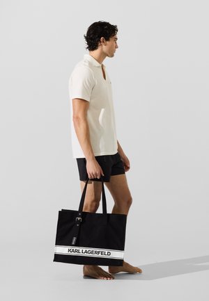 Muškarac u bijelom polo majici i crnim kratkim hlačama nosi veliku crno-bijelu torbu Karl Lagerfeld, stojeći bos na sivoj pozadini.