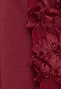 Tissu bordeaux avec une texture lisse d'un côté et une appliqué floral en satin superposé de l'autre, créant un contraste visuel et de la profondeur.