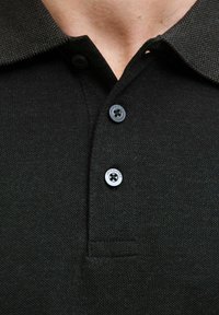 Zwarte polo met een gestructureerde kraag, voorzien van twee grijze knopen met een logo. De stof heeft een geweven patroon en is gedeeltelijk zichtbaar.