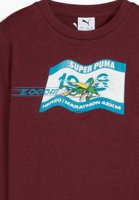 Bordeauxrot T-Shirt aus Baumwolle mit einem grafischen Druck eines grünen Charakters, der läuft, mit blauen Akzenten und dem Text: "SUPER PUMA" und "HERZO/MARATHON 42KM."