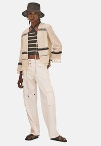 Chaqueta beige corta con ribetes negros y borlas, llevada sobre una camiseta de rayas blancas y negras; combinada con pantalones cargo crema y sandalias marrones.