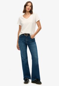 Camiseta blanca de cuello en V combinada con jeans acampanados azules. Los jeans tienen una cintura alta y están adornados con un cinturón negro y hebilla plateada.