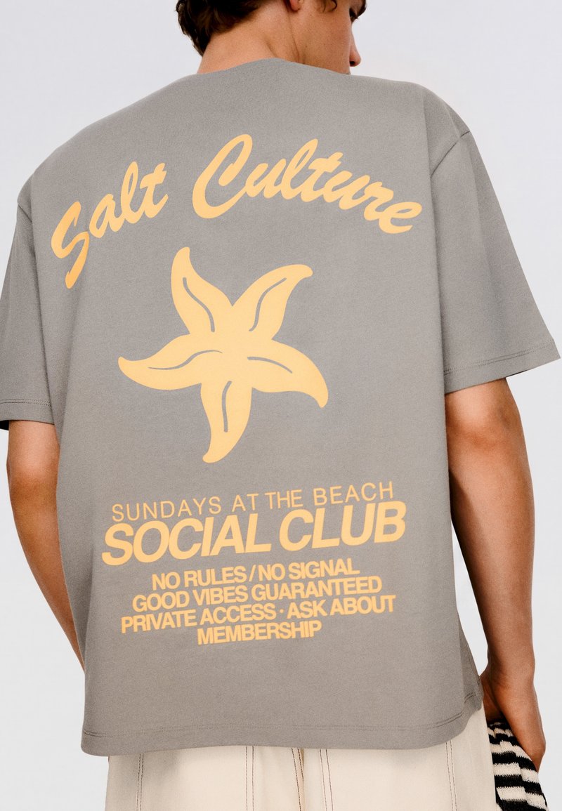 Personne portant un T-shirt gris avec un graphique d'étoile de mer jaune et un texte sur le club social "Salt Culture" et les dimanches à la plage au dos.
