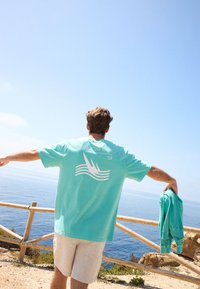 Camiseta de algodón turquesa con diseño gráfico blanco en la espalda, que presenta olas y tres siluetas de aves. Pantalones cortos beige, sosteniendo un suéter azul claro.