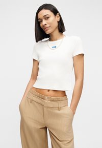 Γυναίκα με ίσια σκούρα μαλλιά που φοράει λευκό κοντό t-shirt, μαργαριταρένιο κολιέ και μπεζ παντελόνι με ρίγες και φαρδιές παντελόνες, με τα χέρια στις τσέπες.